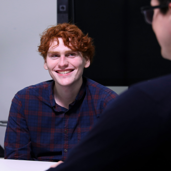 Two people engaged in a conversation; one with red hair smiling while seated across from another person in a dark sweater.